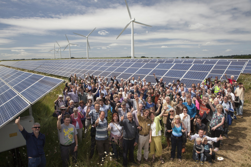 A large group of people giving thumbs up in front of solar panels and wind turbines in a sunny field